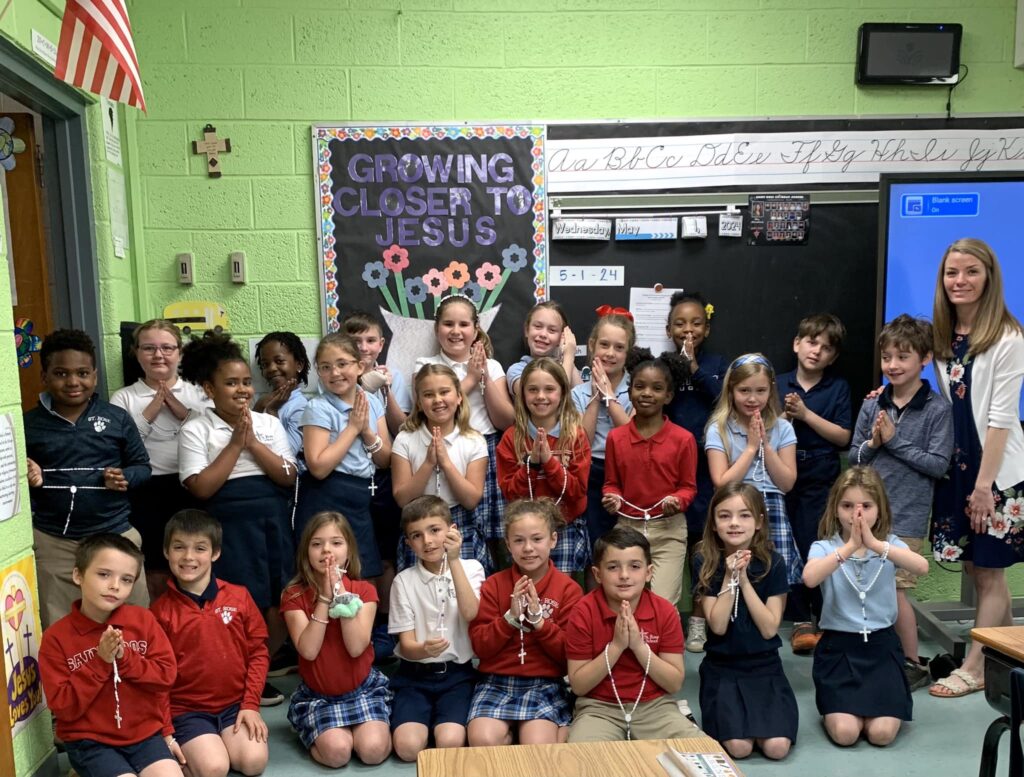 Elementary students and their teacher pose together in a classroom, smiling and holding rosaries with hands folded in prayer in front of a bulletin board that reads ‘Growing Closer to Jesus.’