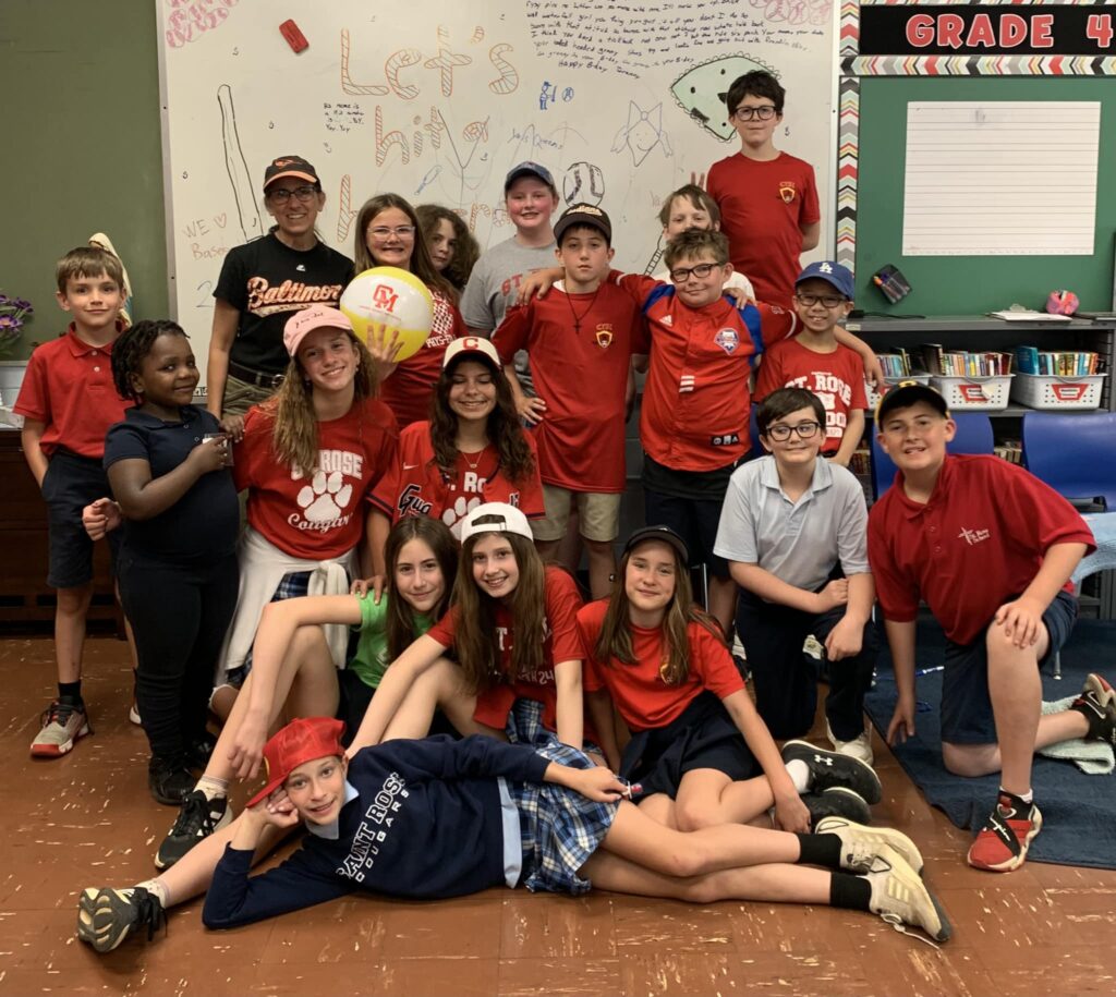 Group of fourth-grade students posing together in a classroom, many wearing red St. Rose shirts and hats, smiling in front of a whiteboard with drawings.
