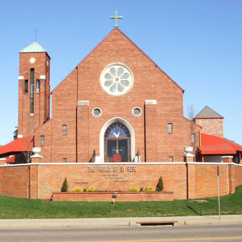 Front view of St. Rose Church, a red brick building with a large rose window, bell tower, and green cross against a clear blue sky.