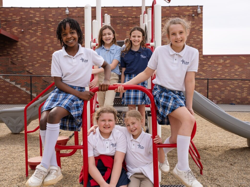 Group of elementary students in school uniforms smiling and posing together on playground equipment at St. Rose Catholic School.