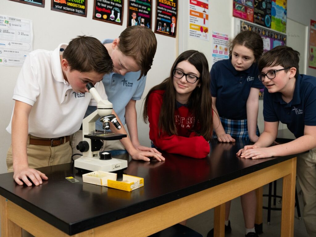 Five students in school uniforms gathered around a science table as one student looks through a microscope, with classmates watching and smiling in a brightly decorated classroom.