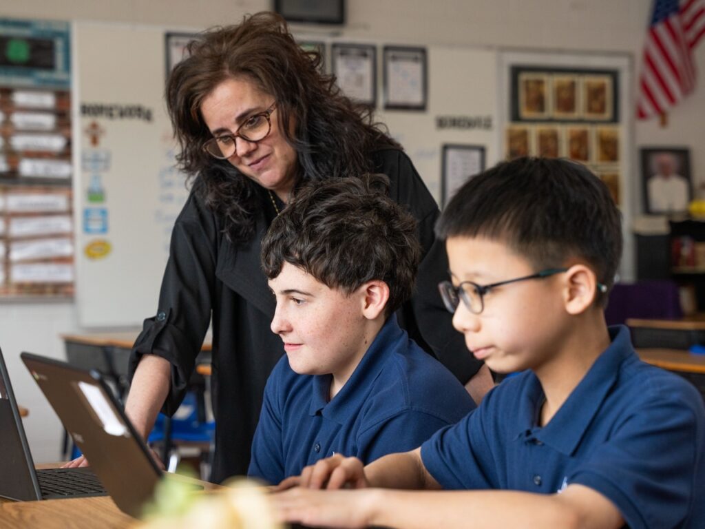 Teacher standing beside two middle school students working on laptops, guiding them during a classroom activity.