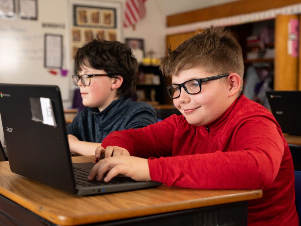 Two elementary students sitting at desks and working on laptops, smiling and focused during a classroom activity.