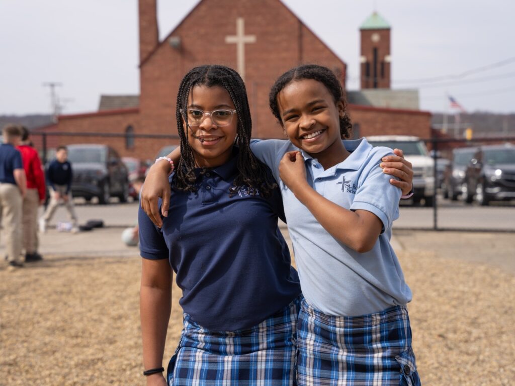 Two smiling students in school uniforms standing together on the playground with their arms around each other, with the school building and other children in the background.