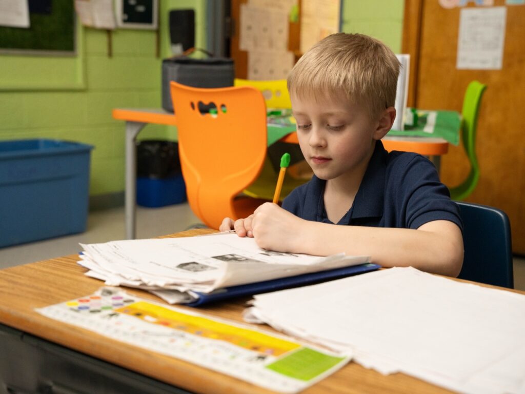 Young student sitting at a classroom desk completing worksheets with a pencil, surrounded by papers and school supplies.