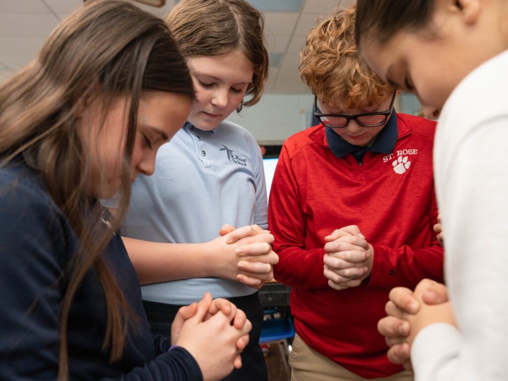 Group of students standing together in a classroom with heads bowed and hands clasped in prayer.