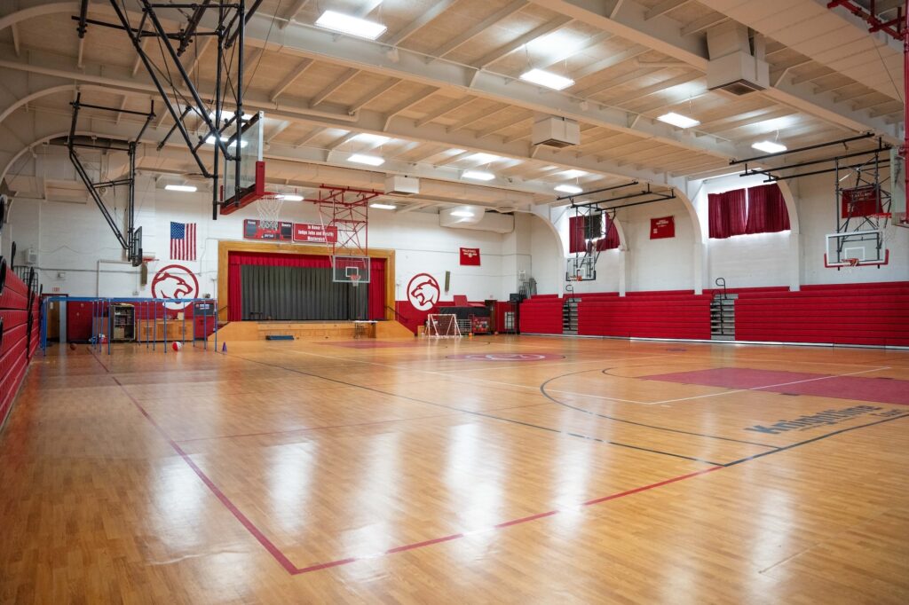 St. Rose Catholic School gymnasium with polished wood floors, red bleachers, multiple basketball hoops, and a stage with red curtains and school mascot graphics on the walls.