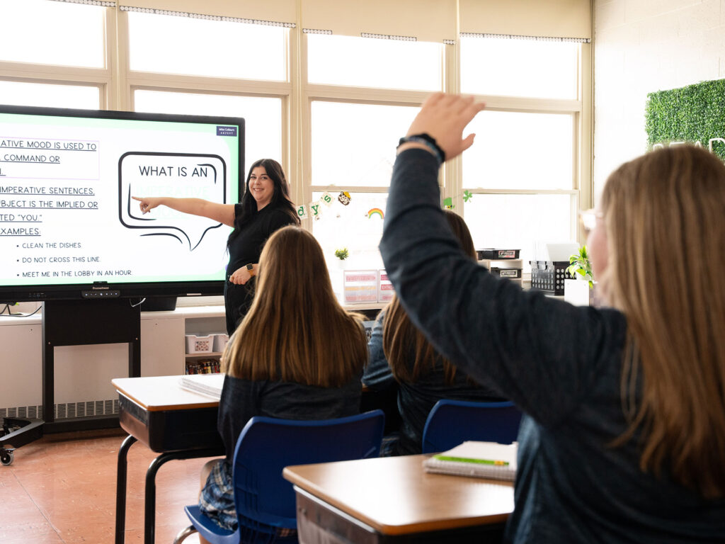 Teacher pointing to a large interactive screen while explaining a lesson on the imperative mood as students sit at desks and one raises her hand to participate.