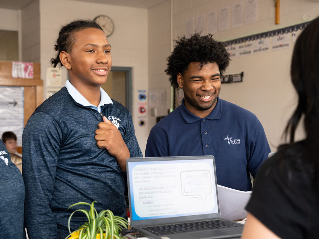 Two high school students smiling and presenting their work on laptops to a teacher in a St. Rose Catholic School classroom.
