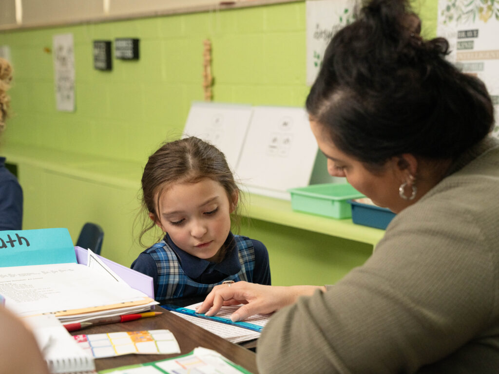 Teacher working one-on-one with a young student in a classroom, guiding her through a math activity at a table with books and worksheets.